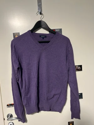 Lila stickad tröja från GANT - Säljer en lila stickad tröja från GANT med v-ringad hals och diskret logga på bröstet. Tröjan har långärmade ärmar och ribbade muddar vid hals, ärmslut och nederkant. Perfekt för dig som gillar klassisk och enkel stil.