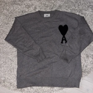 Ljus grå stickad tröja från Ami Paris - Säljer en grå stickad tröja från Ami Paris med svart hjärta och bokstaven A på bröstet. Tröjan har rund hals, ribbade muddar och är långärmad. Perfekt för dig som gillar stilrena detaljer och vill ha något som sticker ut lite. Tröjan är använd 1 gång