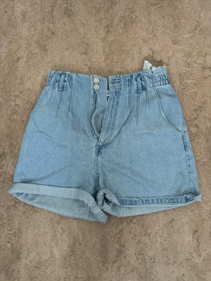 Ljusblå denimshorts från Zara - Säljer ett par ljusblå denimshorts från Zara med hög midja och elastisk bakdel. Shortsen har två framfickor, två bakfickor, uppvikta benslut och stängs med två knappar och dragkedja. Perfekta för varma dagar och chill stil.
