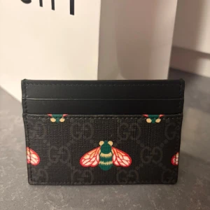 Svart plånbok från Gucci med bi-mönster - Säljer en svart plånbok från Gucci med GG-logga och färgglada bi-motiv. Plånboken har flera kortfack och är tillverkad i skinn med tryckt mönster. Perfekt för dig som vill ha något unikt och snyggt i fickan.