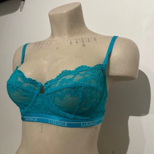 Turkos spets-bh från Lounge - Turkos bh från Lounge med spetsdetaljer och blommigt mönster. Justerbara axelband och elastiskt band med Lounge-logga. Kuporna är genomskinliga med snygg spets och blommor. Ganska säker på att storleken är 80C/D