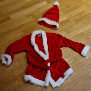 Röd tomtejacka med vit fuskpäls - Röd tomtejacka med vit fuskpäls längs krage, ärmslut och nederkant. Jackan har bälte i midjan och matchande tomteluva med vit kant och tofs. Använt 1 säsong. Jacka och Luva - i fleece. Inga byxor ingår.