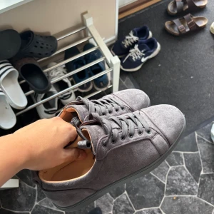Grå mockasneakers från Louboutin - Snygga grå sneakers i mocka från Louboutin med klassiska röda sulor och diskret guldfärgad logga på insidan. Skorna har rund tå, platt sula och grå snörning. Perfekta för dig som vill ha en clean och exklusiv look.
