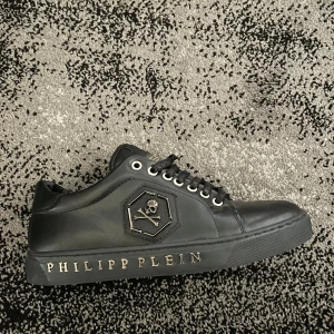 Svarta sneakers från Philipp Plein - Snygga svarta sneakers från Philipp Plein i skinn med cool döskalle-logga på sidan och metallplatta på plösen. Skon har snörning med märkta skosnören och Philipp Plein-text i metall på sulan. Perfekt för dig som gillar exklusiv streetstyle.