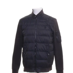 Svart cardigan från Polo Ralph Lauren - Svart cardigan från Polo Ralph Lauren med quiltad front, ribbade muddar och ärmar i mjuk bomullsmix. Jackan har hög krage, dragkedja framtill och fickor med knappstängning. Perfekt för dig som gillar stilren och sportig look. Krage och nedre kant är blek annars bra skick 