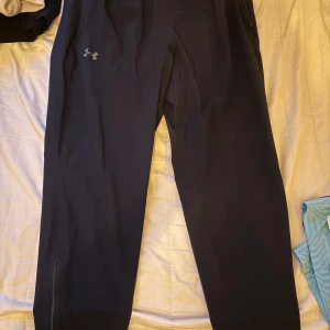 Svarta träningsbyxor från Under Armour XL - Svarta träningsbyxor från Under Armour i storlek XL. Byxorna har en slim passform, diskret logga på låret och är gjorda i ett stretchigt syntetmaterial som andas. Perfekta för löpning eller gym, med dragkedja vid benslut för enkel av- och påtagning.