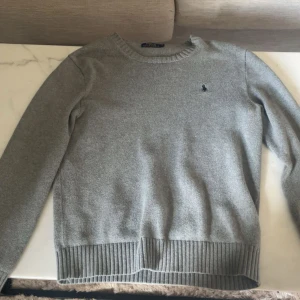 Grå stickad tröja Polo Ralph Lauren - Klassisk grå stickad tröja från Polo Ralph Lauren med rund halsringning och diskret broderad logga på bröstet. Tröjan har ribbade muddar vid ärmslut och nederkant, perfekt för lager-på-lager under kyliga dagar.