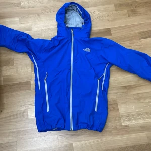 Blå vindjacka från The North Face - Snygg blå vindjacka från The North Face med huva och vita dragkedjor. Jackan har meshfoder, två sidofickor med dragkedja och justerbar huva. Perfekt för blåsiga dagar och outdoor-äventyr. Klassisk logga på bröstet och ryggen.