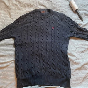 Mörkblå kabelstickad tröja Morris - Kabelstickad mörkblå tröja från Morris med rund hals och diskret röd broderad logga på bröstet. Tröjan har ribbade muddar och är tillverkad i mjuk bomull. Perfekt för lager-på-lager.