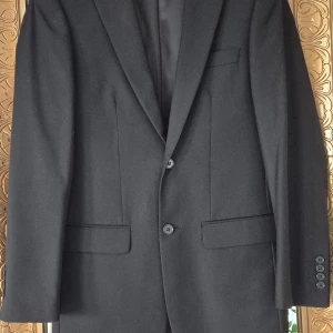 Svart kavaj - Klassisk svart kavaj från Dressmann i storlek 44. Kavajen har två knappar framtill, tre fickor och slits bak. Fodrad insida i blankt tyg och stilren passform.