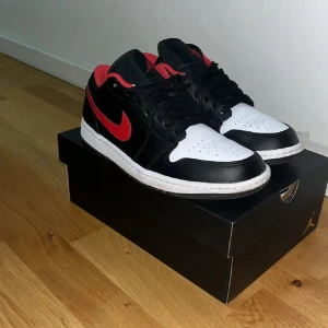 Nike Air Jordan 1 Low svart/röd/vit - Nike Air Jordan 1 Low i svart, vitt och rött skinn. Klassisk siluett med röd Swoosh, svart ovandel, vit tåbox och röd broderad Air Jordan-logga på hälen. Skorna har svarta snören och extra röda snören medföljer. Perfekta för dig som gillar streetwear och sneakers.