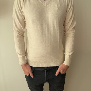  stickad tröja suitsupply - En stilren beige stickad tröja med v-ringning och långa ärmar. Tröjan har ribbade muddar vid ärmslut och nederkant, vilket ger en snygg passform. Perfekt för lager-på-lager och funkar året runt. Från suitsupply