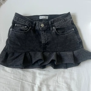 Svart handgjord jeanskjol med volang från Gina  - Säljer en svart handgjord jeanskjol från av ett par Gina jeans med snygg volangkant nertill. Kjolen har klassisk femficksdesign, bälteshällor och knäppning fram. Perfekt för dig som gillar en edgy men söt stil.