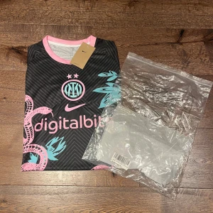 Inter Nike svart fotbollströja rosa orm - Inter fotbollströja från Nike i svart med rosa och turkosa detaljer. Snyggt ormmotiv och växtgrafik framtill, rosa logga och detaljer vid hals och ärmslut. Tillverkad i lätt och ventilerande polyester, perfekt för match eller träning.