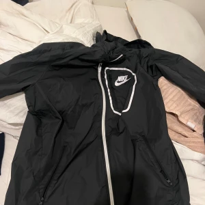 Svart vindjacka från Nike med huva - Svart vindjacka från Nike med vit dragkedja och logga på bröstet. Jackan har huva, två fickor med dragkedja och sportig design. Perfekt för blåsiga dagar och enkel att matcha med streetwear.