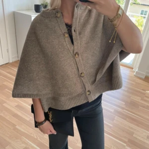 snygg poncho tröja - Snygg poncho tröja med ribbade kanter och stora knappar framtill. Modellen är vid och har en cape-liknande form som ger en chill och trendig vibe. Perfekt att slänga över en långärmad tröja för extra lager. Enkel att styla och supermjuk.