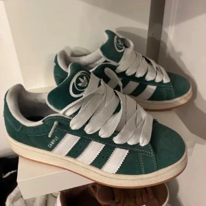Adidas Campus gröna sneakers mocka - Säljer ett par gröna Adidas Campus sneakers i mocka med klassiska vita ränder och breda vita skosnören. Skorna har låg profil, vit sula och Adidas logga på plösen. Perfekta för dig som gillar retrostil och streetwear.