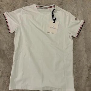 Vit Moncler t-shirt med rödblå detaljer - Vit t-shirt från Moncler i slim fit med klassisk rund hals. Ärmarna har snygga rödblå ränder och Moncler-logga på vänster ärm. Tillverkad i mjuk bomull, perfekt för en clean och stilren look.