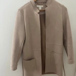 Beige mockaliknande kappa från H&M - Stilren beige kappa från H&M i mockaliknande material. Jackan har raka linjer, stora fickor framtill och ett smalt bälte i midjan. Enkel krage och lång ärm ger en clean look. Perfekt för dig som gillar minimalistisk stil.