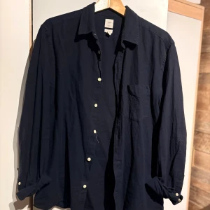 Mörkblå linneskjorta från H&M - Snygg mörkblå skjorta från H&M i luftig linnemix. Klassisk krage, vita knappar och bröstficka. Perfekt oversized fit för en chill och stilren look. Ärmarna kan rullas upp för en avslappnad vibe.