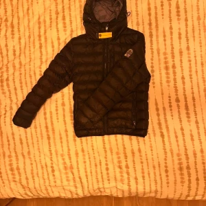 Svart dunjacka från Parajumpers - Svart dunjacka  med huva, gul detalj vid halsen och klassisk. Jackan är superlätt, har dragkedja framtill och två sidofickor med dragkedja. Perfekt för kalla dagar och riktigt snygg streetstil.