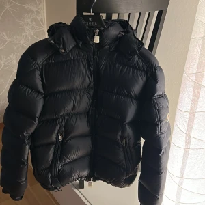 Svart Moncler pufferjacka med huva - Säljer en svart pufferjacka från Moncler med ikonisk logga på ärmen. Jackan har dragkedjor på fickorna, snörning i huvan och en riktigt fet, vadderad look. Perfekt för kalla dagar när du vill hålla stilen och värmen. Klassisk design med hög krage och snygga detaljer. Tveklöst den bästa på Plick just nu med mycket dun. 