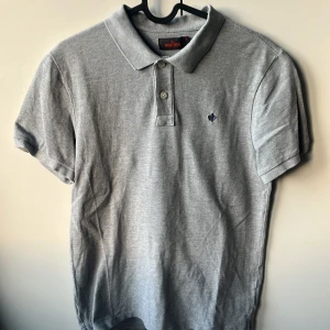 Grå pikétröja från Morris, storlek M passar 170-185 - Snygg grå pikétröja från Morris med klassisk krage och två knappar framtill. Tröjan har korta ärmar och en liten broderad logga på bröstet. Tillverkad i mjuk bomull som känns skön mot huden. Perfekt till jeans.