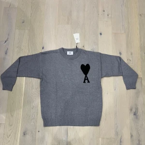 Grå stickad tröja från Ami Paris - Grå stickad tröja från Ami Paris med svart hjärta och bokstaven A broderat på bröstet. Klassisk rund halsringning, ribbade muddar och nederkant. Perfekt för dig som gillar stilren design med en twist.