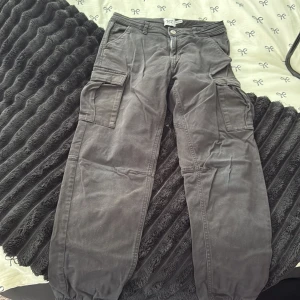 Svarta cargobyxor från 157 - Svarta cargobyxor från 157 med flera stora fickor på sidorna och resår vid bensluten. Byxorna har dragkedja och knapp framtill samt bälteshällor. 