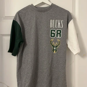 Milwaukee Bucks t-shirt från New Era XS - Grym Milwaukee Bucks t-shirt från New Era i storlek XS. Tröjan är grå med en grön ärm och en vit ärm, tryck med Bucks-logga, nummer 68 och NBA-märke. Perfekt för basketfans som vill sticka ut. Skön bomullskvalitet och snygg passform.
