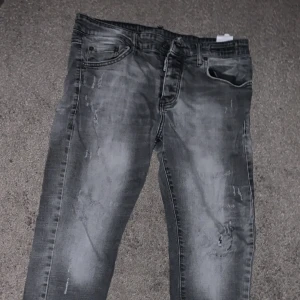 Grå Dsquared2 jeans, storlek 32 - Snygga grå jeans från Dsquared2 i storlek 32. Jeansen har en tvättad look med slitningar och detaljer som ger en cool vibe. Klassisk femficksmodell med dragkedja och knapp. Perfekta för dig som gillar streetstyle och vill sticka ut.