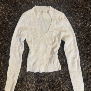 Vit ribbad långärmad topp - Snygg vit ribbad topp med lång ärm och v-ringning samt en cool cut out-detalj vid halsen. Figurnära passform som framhäver siluetten. Perfekt att styla med jeans eller kjol för en trendig look.
