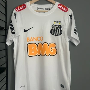 Vit Santos Neymar Jr fotbollströja Nike - Santos hemmatröja med Neymar Jr och nummer 11 på ryggen. Vit med svarta och orangea tryck, klubbmärke på bröstet och flera sponsorloggor. Tillverkad i lätt Dri-FIT-material som andas. Klassisk passform och korta ärmar, perfekt för fotbollsälskare. Helt ny tröja med tagg. En replika 