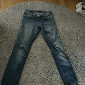 Blå skinny jeans från Nudie Jeans - Säljer ett par klassiska blå skinny jeans från Nudie Jeans med snygga slitningar och kontrastsömmar. Jeansen har fem fickor, normal midja och smal passform. Perfekta för dig som gillar en stilren och modern look.