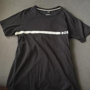 Svart BOSS t-shirt med vit rand - Svart t-shirt från BOSS med vit horisontell rand och logga på bröstet. Tillverkad i ekologisk bomull, slim fit-modell och perfekt för strandhäng. Klassisk rund hals och korta ärmar.
