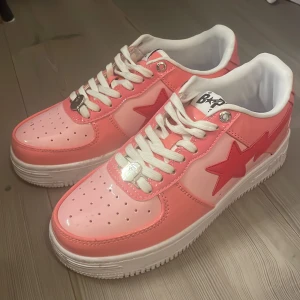 BAPE STA sneakers i rosa  - Säljer ett par BAPE STA sneakers i blankt rosa och rött med vit sula och vita skosnören. Skorna har den ikoniska stjärnan på sidan och BAPE-logga på plösen. Perfekta för dig som vill sticka ut med färg och streetstyle.
