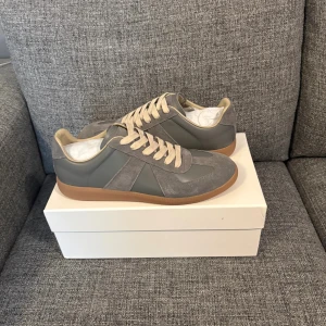 Maison margiela - Snygga grå sneakers med beige snörning och detaljer i mocka. Skorna har en klassisk siluett med rund tå och platt gummisula i brunt. Perfekta för dig som gillar stilrena och neutrala färger. Passar till det mesta och är riktigt bekväma.