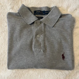 Grå pikétröja från Polo Ralph Lauren - Klassisk grå pikétröja från Polo Ralph Lauren med broderad logga i vinrött på bröstet. Tröjan har korta ärmar, krage och knäppning med två knappar. Tillverkad i mjuk bomull och har en normal passform. Perfekt för en clean och stilren look.