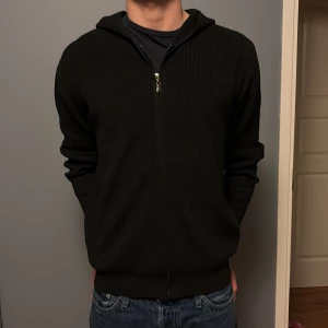 Svart stickad hoodie med dragkedja - Svart stickad hoodie med dragkedja framtill och huva. Tröjan har långärmad passform och är ribbad i materialet, vilket ger en snygg struktur. Perfekt för dig som gillar en enkel och stilren look.