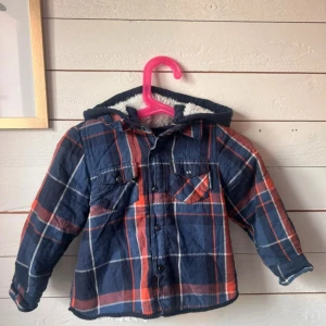 Rutig overshirt med teddyfoder och huva - Snygg rutig overshirt med blå, röda och vita toner. Jackan har huva och är fodrad med mjukt, vitt teddyfoder på insidan. Två bröstfickor med knappar och klassisk skjortkrage. Perfekt för kyliga dagar när du vill vara både varm och stilren.