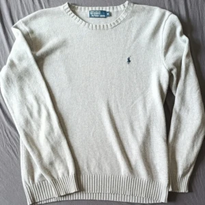 grå stickad tröja från Polo Ralph Lauren - Klassisk beige/grå stickad tröja från Polo Ralph Lauren med rund halsringning och broderad marinblå logga på bröstet. Tröjan har ribbade muddar vid ärmslut och nederkant, perfekt för lager-på-lager under kyliga dagar.