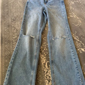Blå bootcut jeans med slitna knän - Säljer ett par ljusblå bootcut jeans med slitna detaljer vid knäna. Jeansen har klassisk femficksdesign och är tillverkade i jeansmaterial. Perfekta för en avslappnad och trendig look.