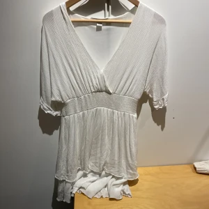 Vit omlottklänning från Amisu - Supersöt vit kortklänning från Amisu med omlott och v-ringning. Klänningen har korta ärmar, markerad midja och lätt plisserat tyg som ger en flowy känsla. Perfekt för sommarens alla tillfällen och enkel att styla med accessoarer.