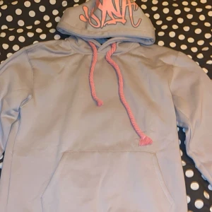 Grå synaworld hoodie  - Säljer en grå hoodie från synaworld med rosa snören och tryck på huvan. Den är oanvänd och i storlek M.  Kan göra Meet ups, endast i stockholm, helst city.