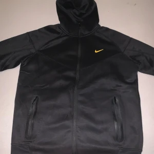 Svart Nike Nocta  - Nike NOCTA hoodie i svart med gul Swoosh. Exklusiv design, premiumkvalitet och praktiska fickor med dragkedja. Ett eftertraktat plagg för dig som vill ha både stil och komfort.