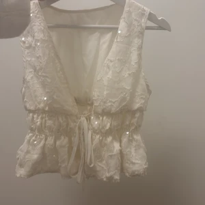 Vit omlottopp med paljetter och knyt - Supersöt vit omlottopp med v-ringning, knyt i midjan och glittriga paljettdetaljer. Toppens tyg har en lätt strukturerad yta med broderade inslag och volangliknande kant nedtill. Perfekt för dig som gillar romantiska och unika plagg.