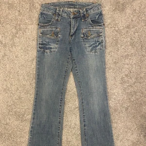 Blå bootcut jeans med fickdetaljer - Snygga blå jeans med bootcut passform och coola fickor med lock och knapp både fram och bak. Jeansen har klassisk tvätt och kontrastsömmar, perfekt för dig som gillar en avslappnad men trendig stil. Midja rakt över: 33cm Lår: 21cm Innerben: 37cm