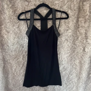 Svart linnetopp med breda nitaxelband - Snyggt svart linne med breda axelband dekorerade med silverfärgade nitar. Toppen har en unik rygg med korsade band.