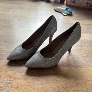 Glittriga silverfärgade pumps - Snygga pumps från Tamaris i glittrigt silvertyg med spetsig tå och smal klack. Perfekta för dig som vill sticka ut med en elegant och trendig look. Skorna har en klassisk form och är täckta med ett skimrande material som fångar ljuset.