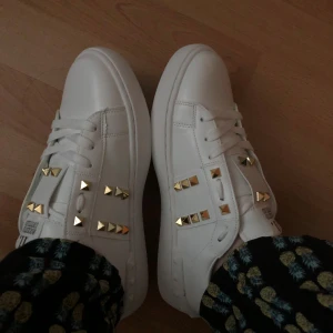 Valentino Rockstud vita sneakers - Snygga vita Valentino Garavani Rockstud sneakers i skinn med ikoniska guldiga nitar på sidorna och tungan. Klassisk rund tå, platt sula och coola detaljer längs yttersulan. Perfekta för dig som vill sticka ut med en lyxig touch.